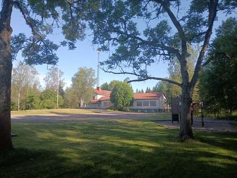 Vanha punakattoinen koulurakennus, jonka ympärillä on vehreä piha. Lehtipuut varjostavat rakennuksen pihanurmea.