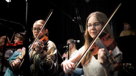 I förgrunden spelar två studerande fiol, i bakgrunden syns andra musiker.