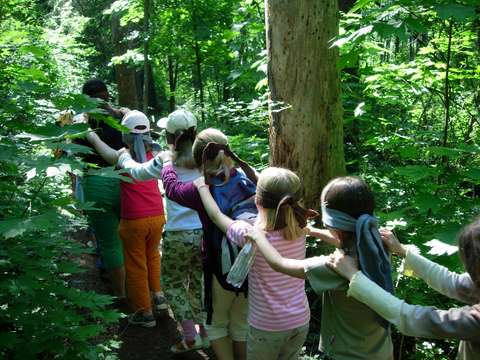 Barn står i ett tåg på en naturstig. Barnen har bindel för ögonen och händerna på axlarna på den som går framför.