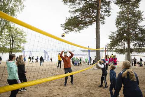 Kuvassa joukko aikuisia ja lapsia pelaa rantalentopallon rannalla. Taustalla näkyy muita ihmisiä.