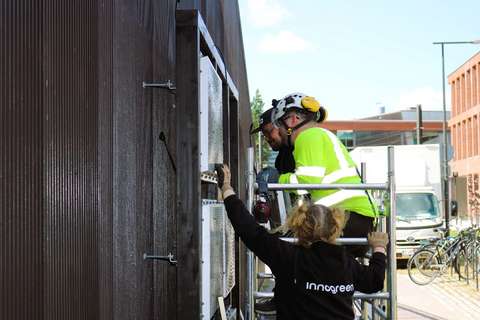 Tre människor i arbetskläder på en byggnadsställning fäster väggens stålelement på en trästomme.