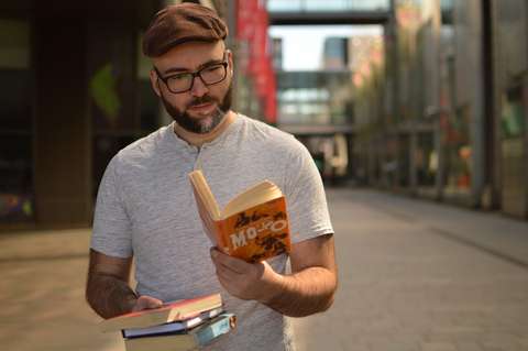 An adult is standing outside reading a book. In addition to the book they are reading, the person is carrying other books.