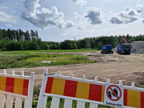Hiekkatietä, kaksi kuorma-autoa ja kaivuri sekä sorakasa ja nurmialuetta. Taustalla puita ja pilviä taivaalla.