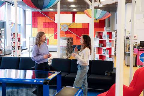 Two women are standing and talking in a colorful library room.