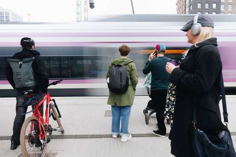 Takaapäin kuvattuja ihmisiä seisoskelemassa junalaiturilla. Taustalla näkyy ohiajava lähijuna.