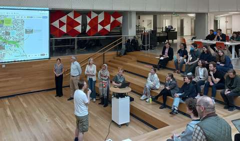 Audotoriotilassa noin 20 henkilöä seuraa kun puhuja esittelee valkokankaalle heijastettua julistetta. 