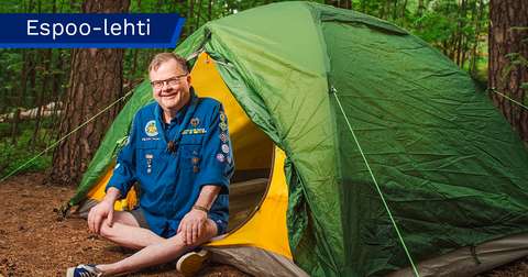 Timo Muttonen, dressed in a scout uniform, is sitting on the ground in front of a tent.