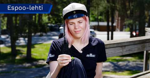 Apila Huhtinen is wearing a student cap, sitting on a bench and looking at a clover she is holding in her hands.