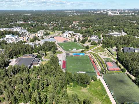 Ilmakuva, jossa näkyy rakennuksia, jalkapallokenttiä ja metsikköä.