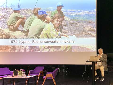 Luennoitsija puhuu mikrofoniin ja taustalla valkokankaalla vanha valokuva.