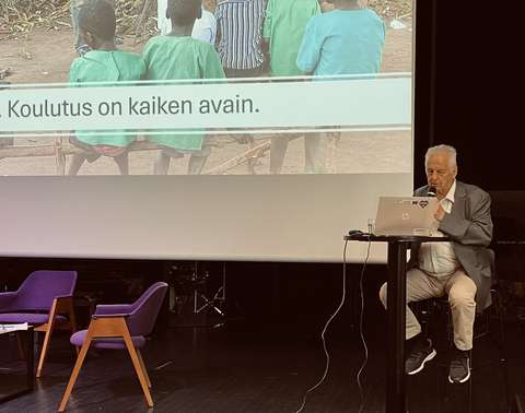 Luennoitsija seisoo auditorion lavalla ja puhuu mikrofoniin.