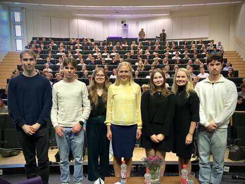 Seitsemän henkilöä rivissä auditorion lavalla yleisön edessä.