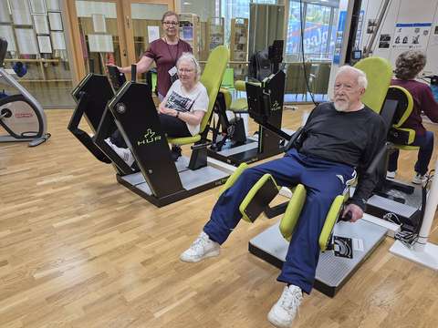 Kuvassa nainen seisoo kuntosalilaitteita käyttävien senioreiden vieressä.