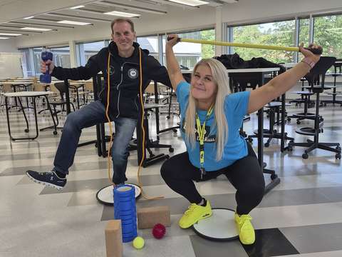 In the picture, a woman and a man are balancing on balance boards in a classroom, holding various pieces of exercise equipment in their hands.