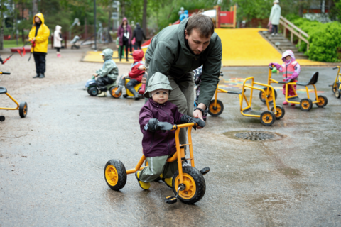 Isä auttaa pientä lastaan pyöräilemään asukaspuistossa.