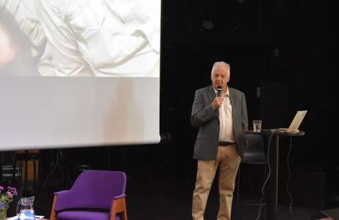 Luennoitsija seisoo auditorion lavalla ja puhuu mikrofoniin.