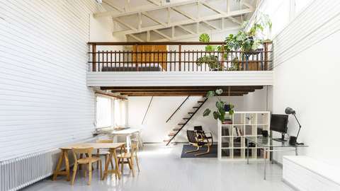 Kuva valoisasta ateljeeasunnosta, jossa etualalla pöytäryhmä ja kirjahylly sekä parvi, jolla kasvaa viherkasveja.