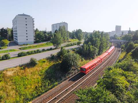Maan päällä kulkeva metrojuna ja raiteet. Ympärillä pensaita, puita, autotie ja muutama kerrostalo.