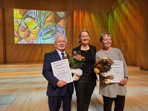 Mies ja kaksi naista seisovat vierekkäin hymyillen. Kuvassa ovat Olli Männikkö Espoon vanhusneuvostosta, Espoon kaupunginhallituksen puheenjohtaja Mervi Katainen ja vuoden eläkeläiseksi valittu Marja Koskenalho. Takana näkyy värikäs taulu ja tyhjää huonetta valaistuna eri värisillä valoilla.