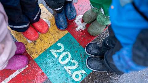 Pienessä ympyrässä seisovien lasten kumisaappaat muodostavat ympyrän ja maassa on vuosiluku 2026.