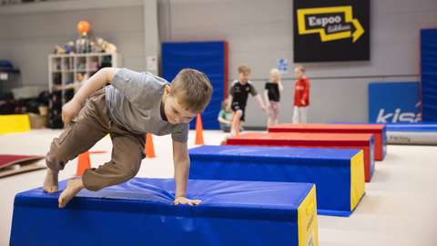 Bilden visar några barn i en sporthall. Det finns några mjuka hinder på golvet. En av barnen hoppar över hindren. De andra barnen står och väntar på sin tur. På väggen ser man en Espoo liikkuu -logga.