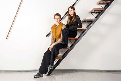 Två personer sitter på trappan till en lägenhetsbyggnad och tittar in i kameran.