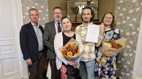 Kuvassa neljä ihmistä kahdessa rivissä. Kolme miestä ja nainen seisovat takarivissä, ja heidän edessään keskellä seisoo nainen. Naisilla kukkakimput kädessä, yhdellä miehistä kehystetty kunniakirja. Takana tapettia ja ovi.