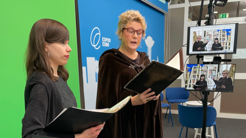 Inari Mykkänen ja Pirkko Ilmanen seisovat studiossa greenscreenin edessä. Sivulla olevalla näytöllä näkyvät esiintyjät taustallaan kirjahylly.