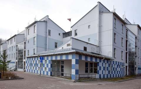 Kiinteistö Oy Kylänaukio kuvattuna pilvisellä ilmalla. Rakennuksessa on sinivalkoinen ruudullinen seinä ja muoto ei ole perinteinen kerrostalo.