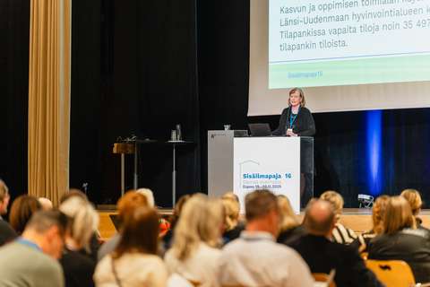 "Toimitusjohtaja Maija Lehtinen seisoo lavalla ja pitää puhetta yleisölle, taustalla näkyy Lehtisen esittämä materiaali..