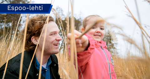 Pieni lapsi seisoo kaislikossa ja osoittaa kaislaa kameralle hymyillen. Vieressä mies istuu kyykyssä katsoen lasta hymyillen.