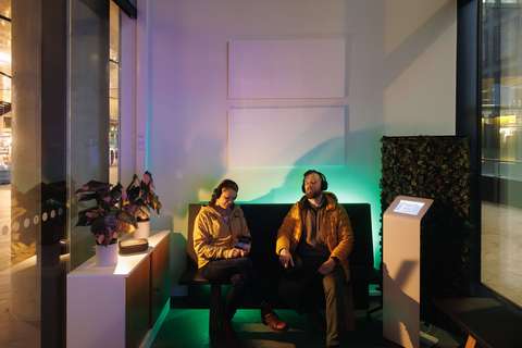 Kaksi ihmistä istuvat penkillä tunnelmallisesti valaistussa hämärässä odotustilassa. Molemmilla on päässä mustat kuulokkeet, ja heidän taustallaan seinällä on kaksi valkoista akustiikkapaneelia.