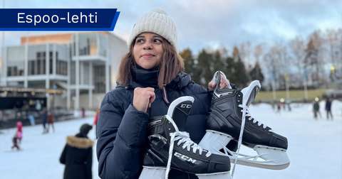 står på en uterink i ett vinterlandskap och håller ett par svarta skridskor i händerna. I bakgrunden syns andra skridskoåkare samt Esbo kulturcentrum.