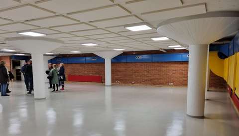 Iso, matala ja tyhjä hallimainen tila, jossa seinät tiiltä ja osittain  maalattu eri väreillä. Kolme iso pylvästä, yhden takana seisoo joukko ihmisiä.