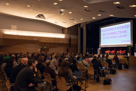 Isossa salissa ihmisiä istuu tuoliriveillä kuuntelemassa puhujaa lavalla. Puhujan taakse lavalle on heijastettu esitys.