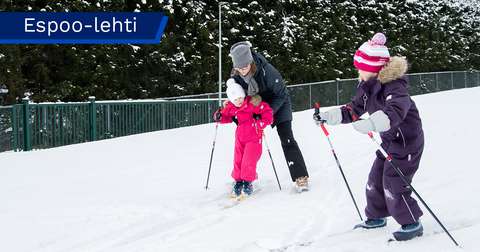 En vuxen och två barn åker skidor på ett snötäckt spår i ett vinterlandskap. Den vuxna hjälper det yngre barnet, medan det äldre barnet åker strax bredvid.