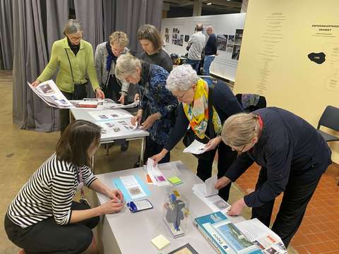 Sex personer står runt ett bord och tittar på bilder, en person sitter på huk på golvet och hjälper till.