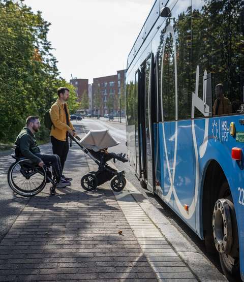 Henkilö, joka istuu pyörätuolissa ja toinen henkilö, joka työntää lastenrattaita ovat menossa sinisen bussin kyytiin bussipysäkiltä.