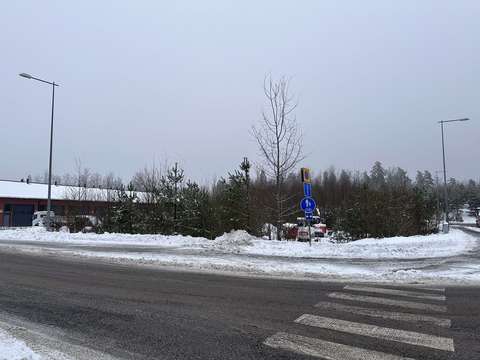 Rakentamaton tontti, jossa kasvaa puita ja säilytetään työmaakoneita. Tontin vieressä on risteys ja suojatie. Tontin toisella puolella on varastorakennus.