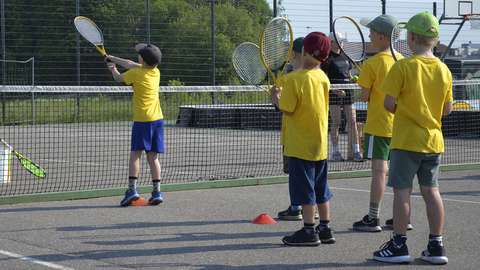 Kuvassa on viisi lasta pelaamassa tennistä ulkokentällä.