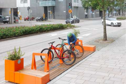 Ruskeaa puuta ja oranssia metallia yhdistelevä pyöräpysäkkirakenne kaupunkimaisemassa. 