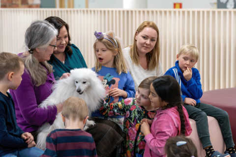Leppävaaran koulun rehtori, Monikon päiväkodin johtaja sekä Monikon päiväkodin eskariopettaja sekä eskarilaisia istumassa sohvalla ja silittämässä koiraa.