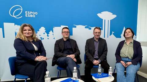 Neljä henkilöä istuu. Taustalla piirretty siluetti Espoon kaupungista.