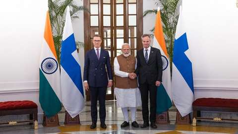 Three men stand and pose for the camera between large Indian and Finnish flags.