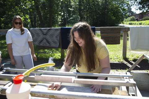 Henkilö pesee mattoa matonpesupaikalla. Takana toinen henkilö ja kuivumassa olevia pestyä mattoja.