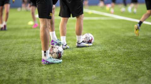 Bilden visar ett konstgräsplan i en hall. Det är unga som spelar fotboll på planen.