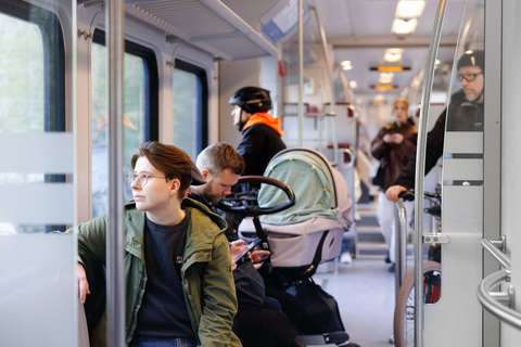 Ihmisiä lähijunassa katselemassa ulos ikkunasta. Yhdellä matkustajalla on mukana pyörä, toisella lastenrattaat.