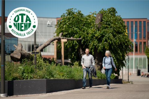 Kaksi ihmistä kävelemässä kesäisessä Otaniemessä Väreen edustalla. Logo: Otaniemi kestävällä tiellä, toukokuu 2026.