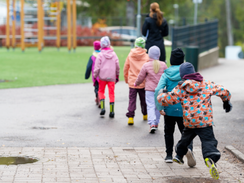 Espoolaiset koululaiset kulkevat jonossa koulun pihalla opettajan perässä. Jonon viimeinen hyppää iloisesti vesilammikon yli. 