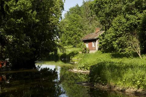 En röd stuga vid en å, omgiven av frodiga lövträd.